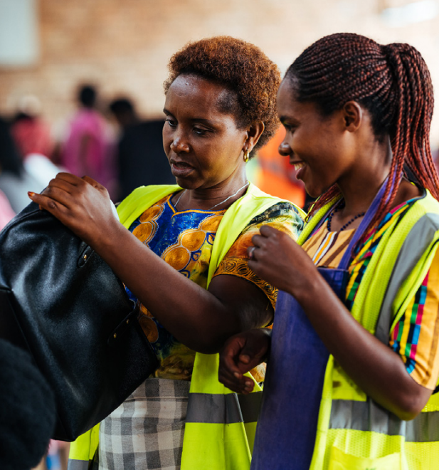 A person inspecting a handbag as another smiles, looking on.