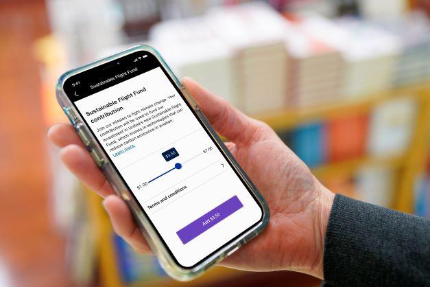 A hand holding a cell phone open to show the screen "Sustainable Flight Fund Contribution" and a sliding scale showing amount contributed.