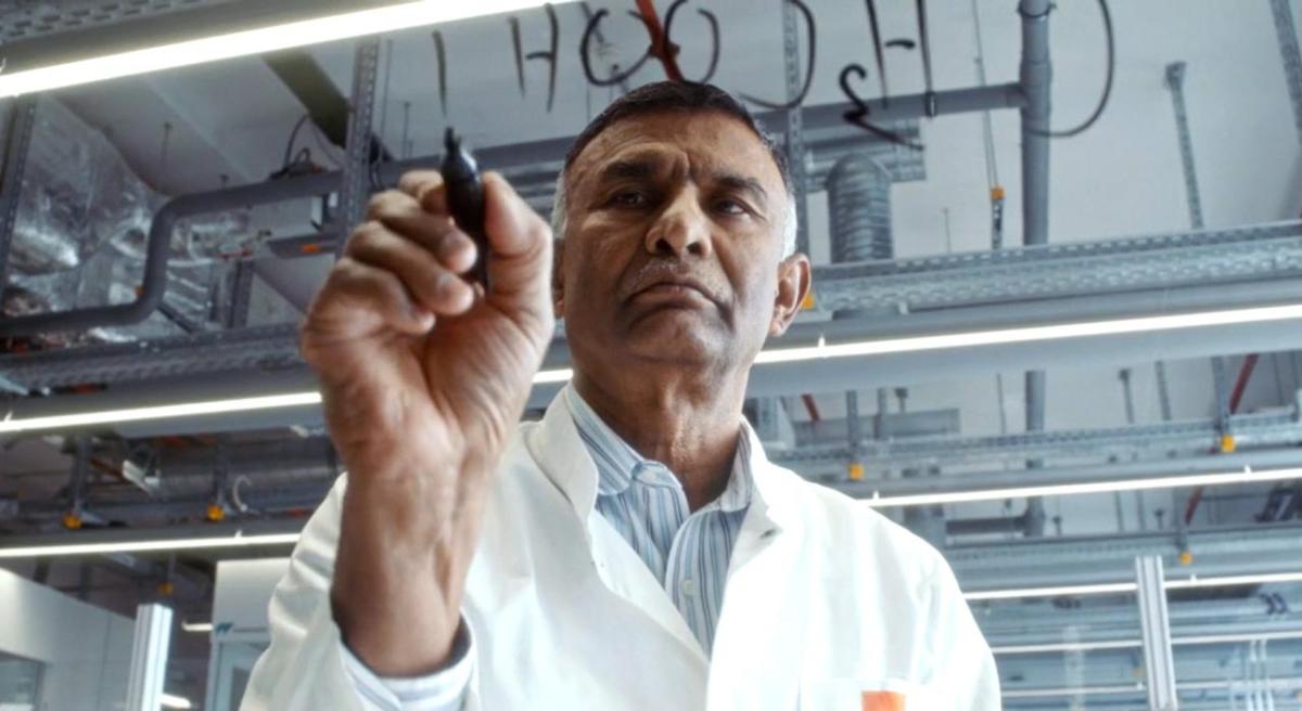 Dr. Ponnusamy wears a lab coat writing out chemical compounds on glass with a marker.