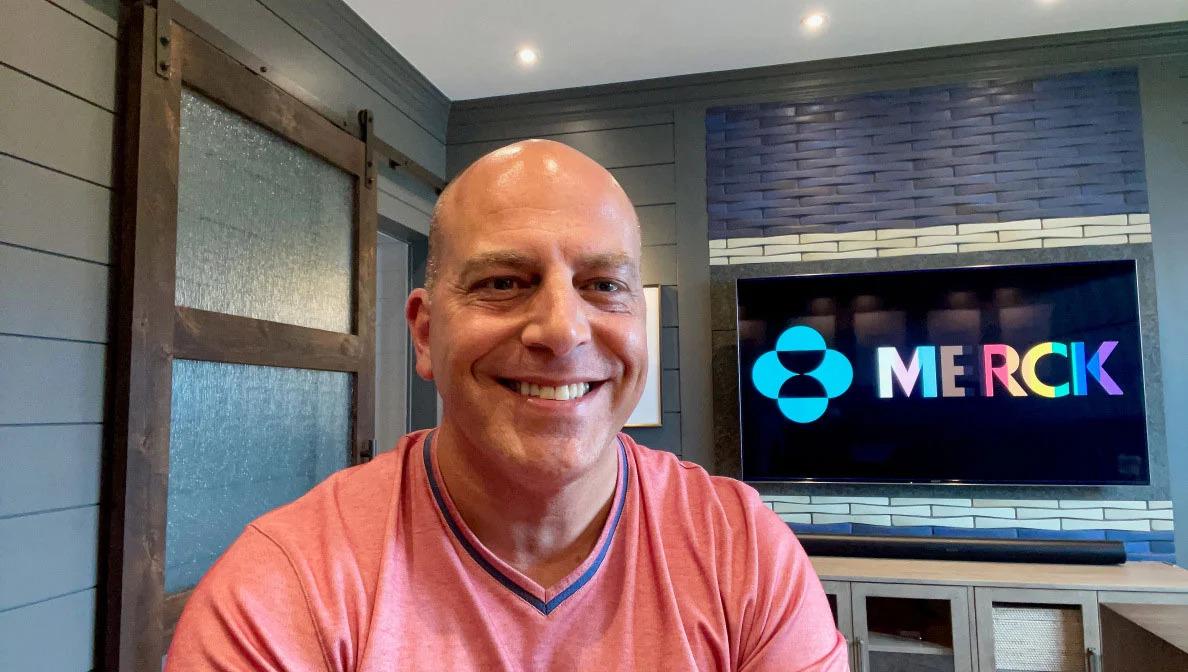 scott wright with a screen behind him playing the Merck logo in LGBTQ+ colors