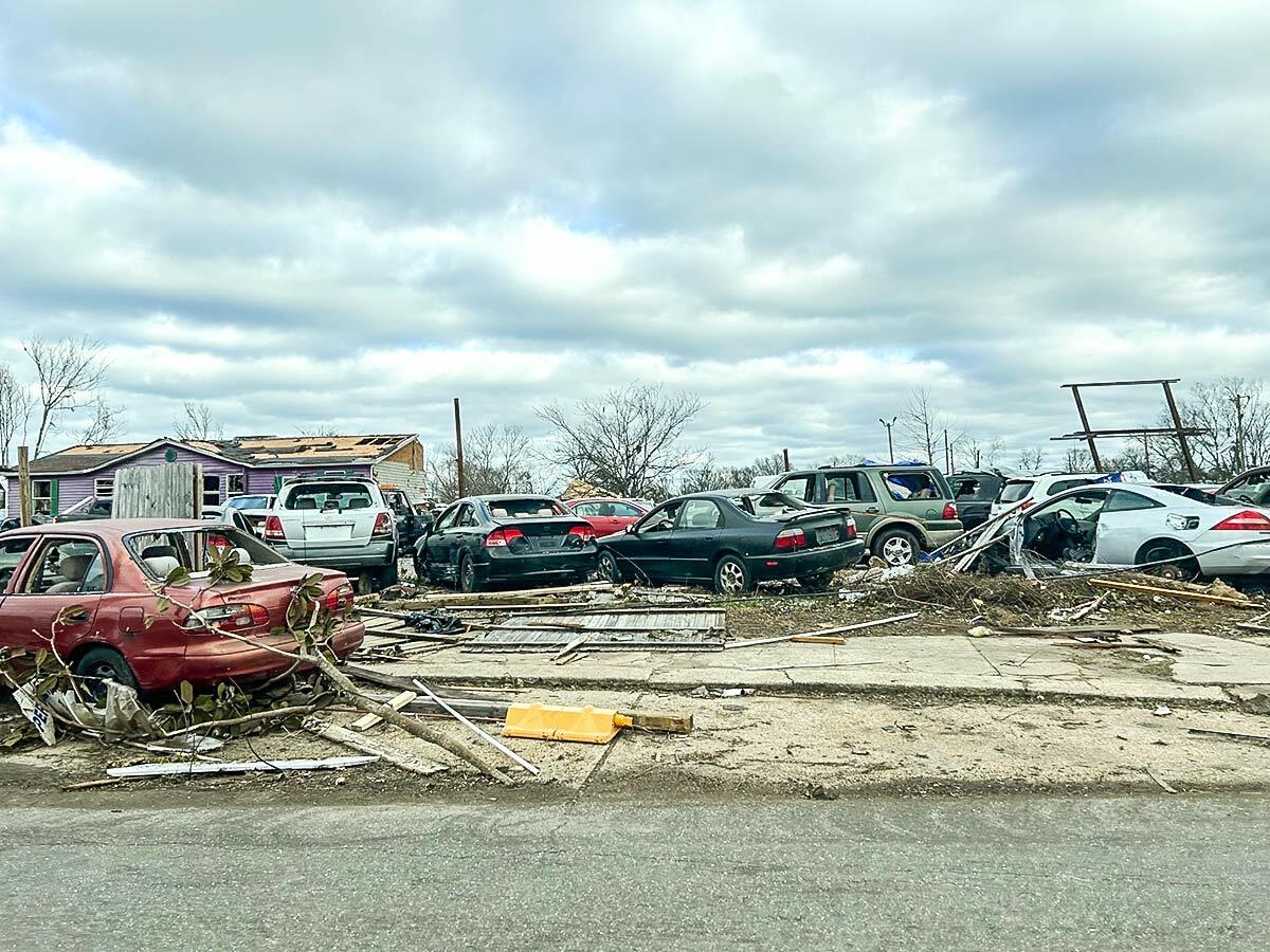 A group of damaged cars and building, debris has broken windows and is strewn around the street and lot.