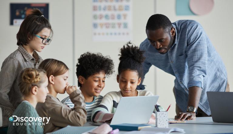 A teacher looking over students who are sat at a table working on a laptop