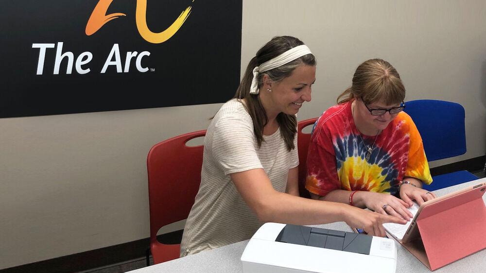 a person in a white t-shirt sits next to a person in a tie-dyed t-shirt, both looking at a laptop and smiling