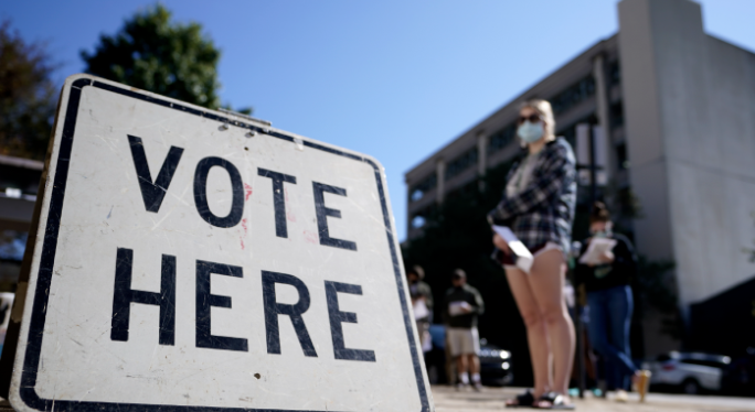 Woman stands if front of a Vote Here sign.