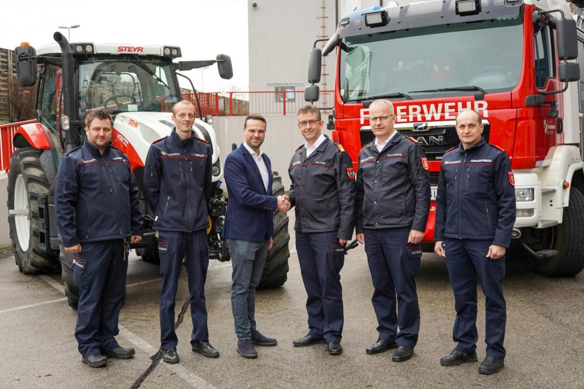 Six people stand in a line, the middle two shake hands. A fire truck and tractor behind them.