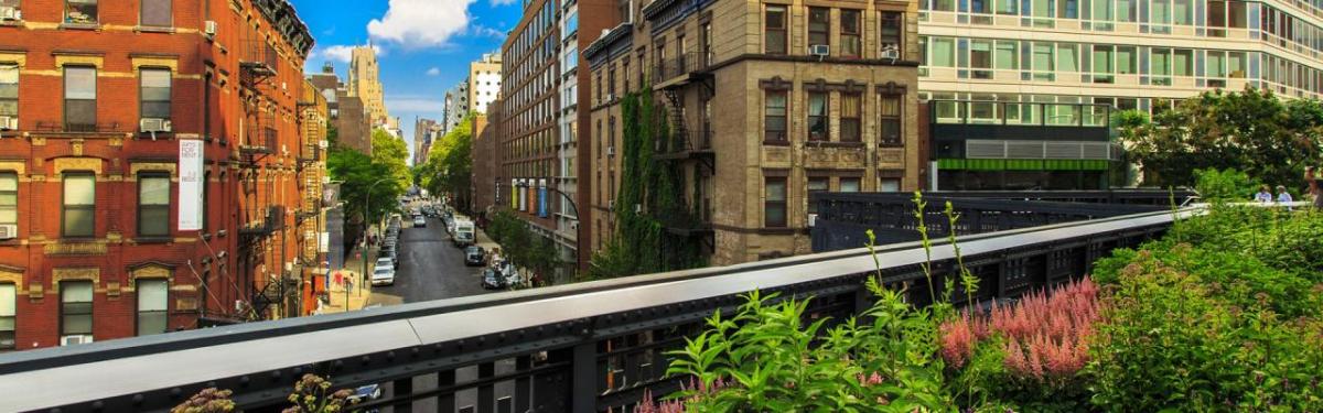 Panoramic view of an urban city with tall, close buildings. An elevated train line running through it. Greenery on the side.