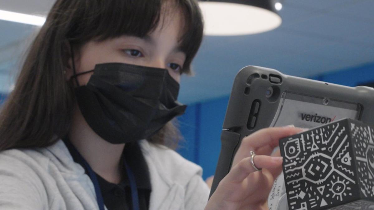 A person wearing a protective mask looks at a verizon tablet and holds a puzzle box