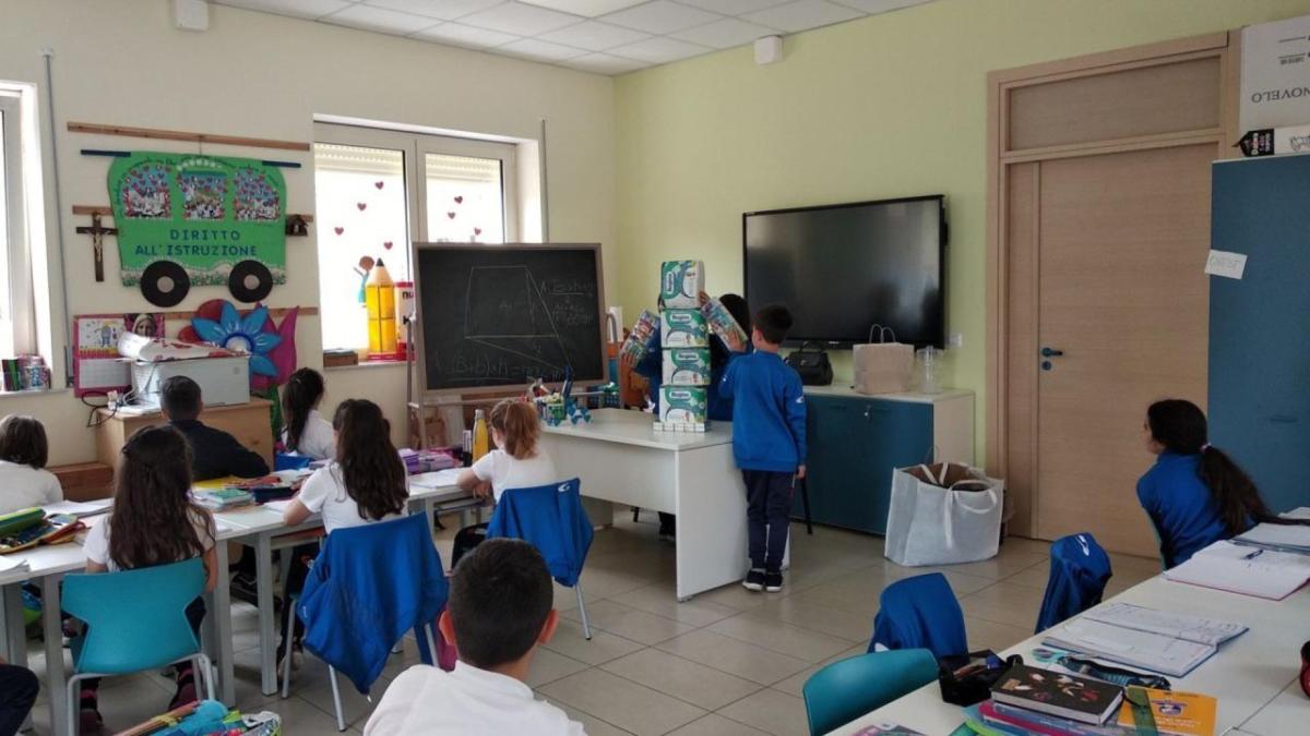 A room of children and a desk at the front stacked with paper products.