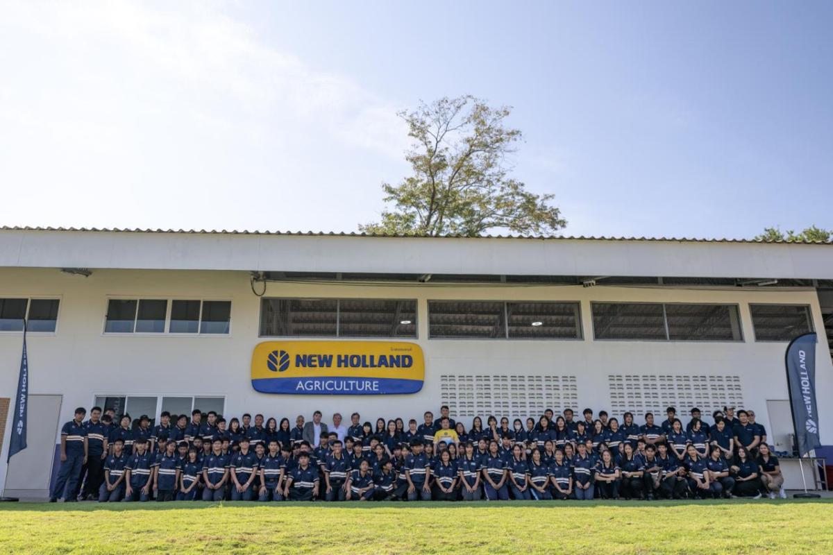 A large group posed outside a long building "New Holland" sign on the wall behind them.