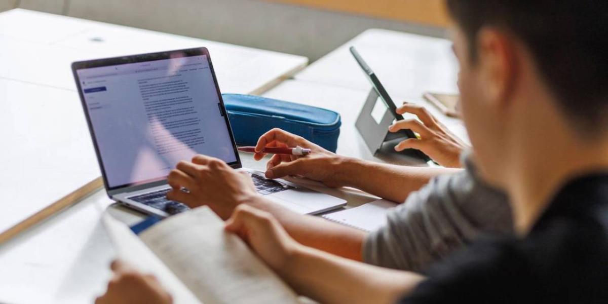 Looking over the shoulder of a person with an open book, another to their right using a laptop on a desk.