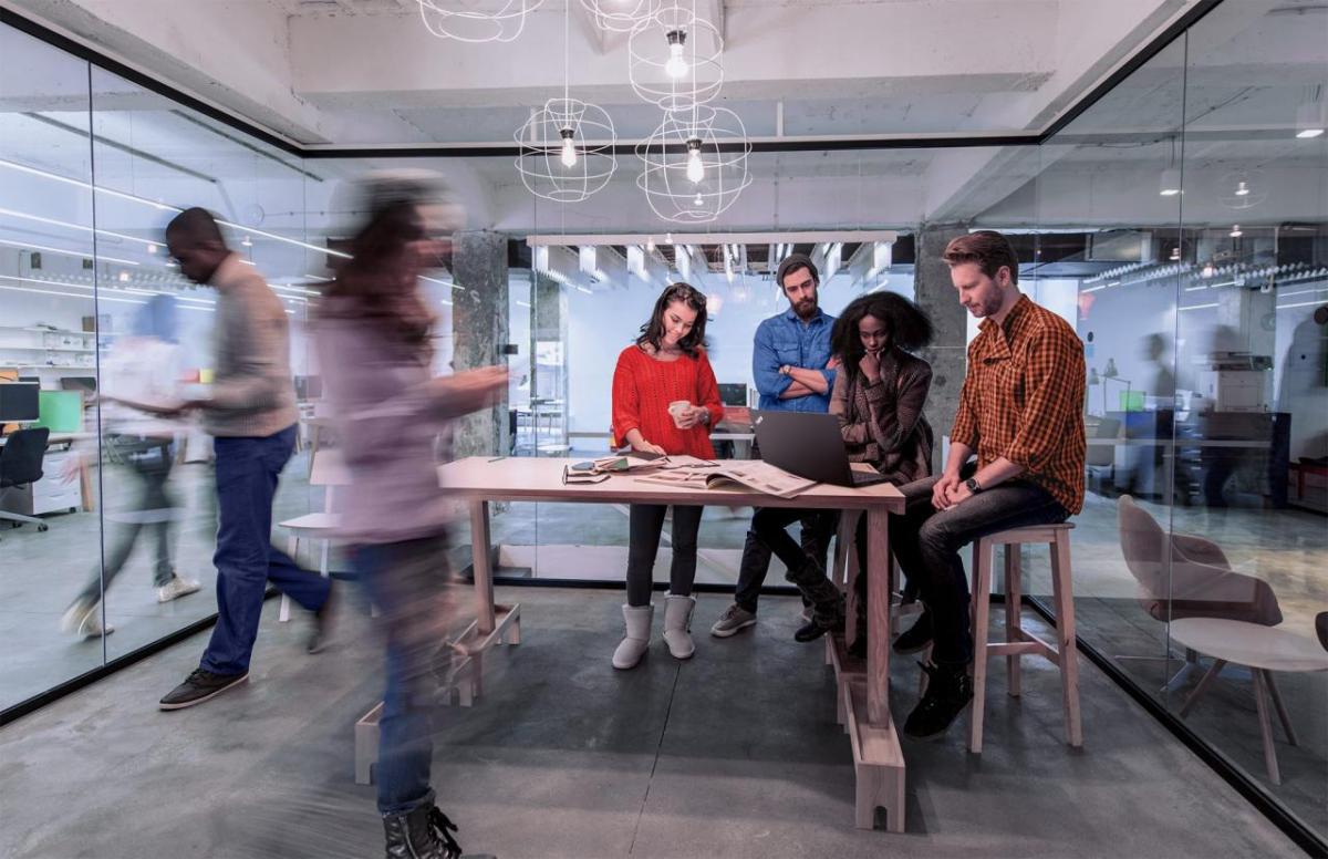 Four people looking at a laptop on a tall desk. Blurred others going by in a busy office setting.