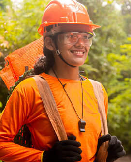 A smiling person in bright orange and safety gear in a forest setting.