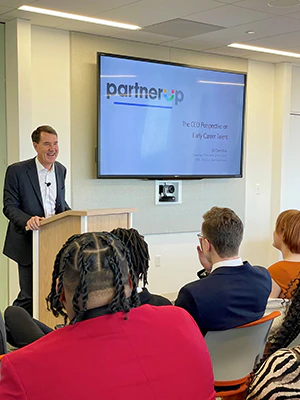 William Demchak standing at a podium speaking to a room of students and adults. A screen behind him displays "Partner up"