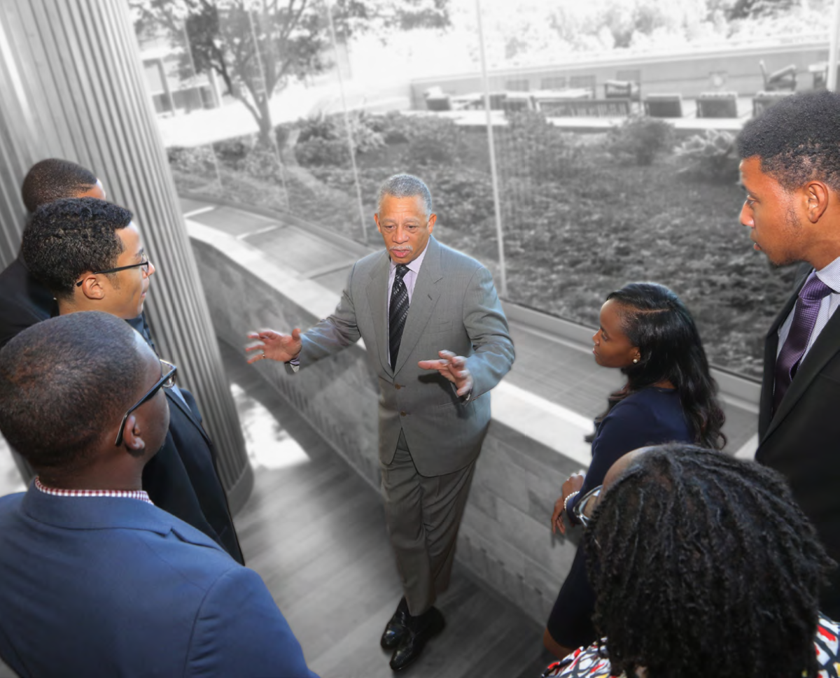 a group of people gather around one speaker in front of a wall of windows