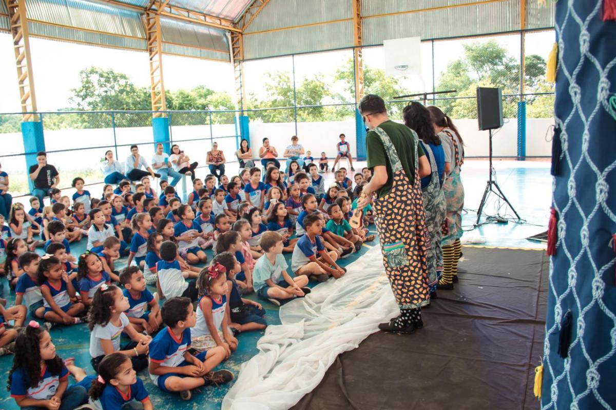 People in costumes stand in front of a large group of seated children in an open building.