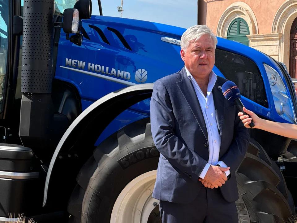 Carlo Lambro in front of a blue New Holland tractor, another holding a microphone for them. 