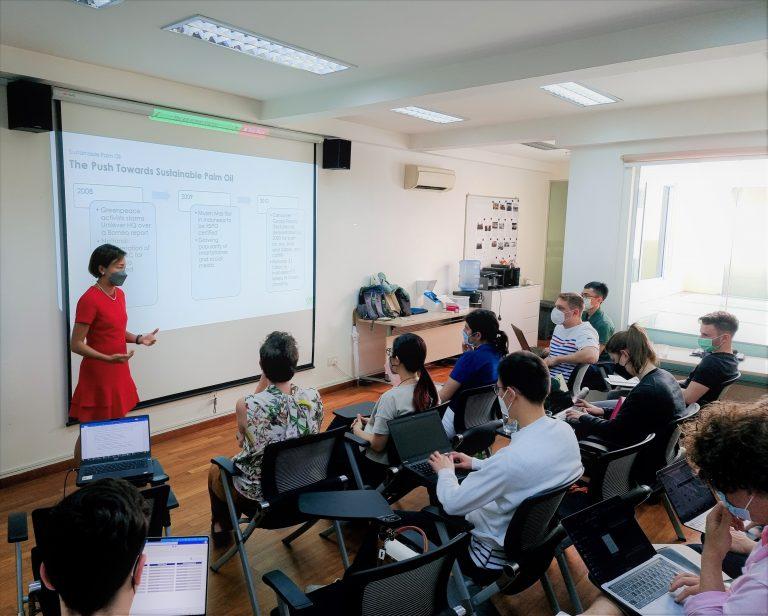 one person stands in front of a small classroom in front of a screen with a presentation being projected on it