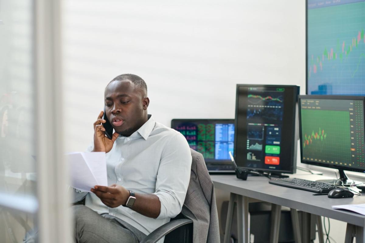 A person talking on a phone, looking at papers, their back to multiple computer screens.