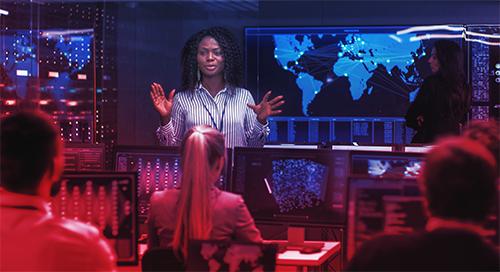 A person standing in front of others seated behind computer screens. A digital world map on the wall behind them.