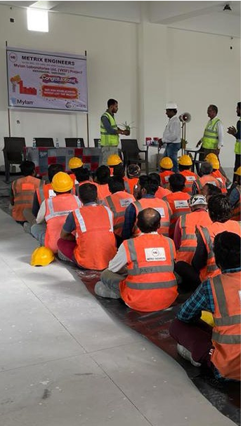 A group seated on the floor, each wearing high-vis vests and some in hard hats. People at the front of the room speaking to them.