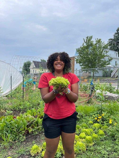 A smiling person holding a bundle of produce outside in a garden.