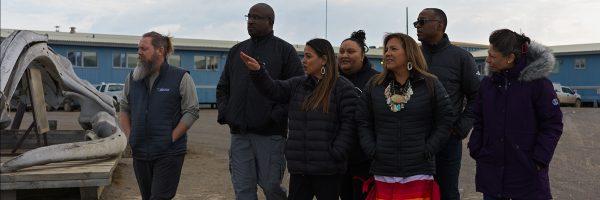 A group gathered outside looking in the same direction. One pointing. A blue long building behind them.