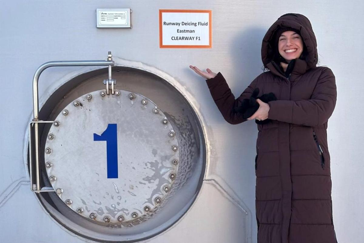 A smiling person pointing to a tank with a sign "Runway deicing fluid. Eastman."