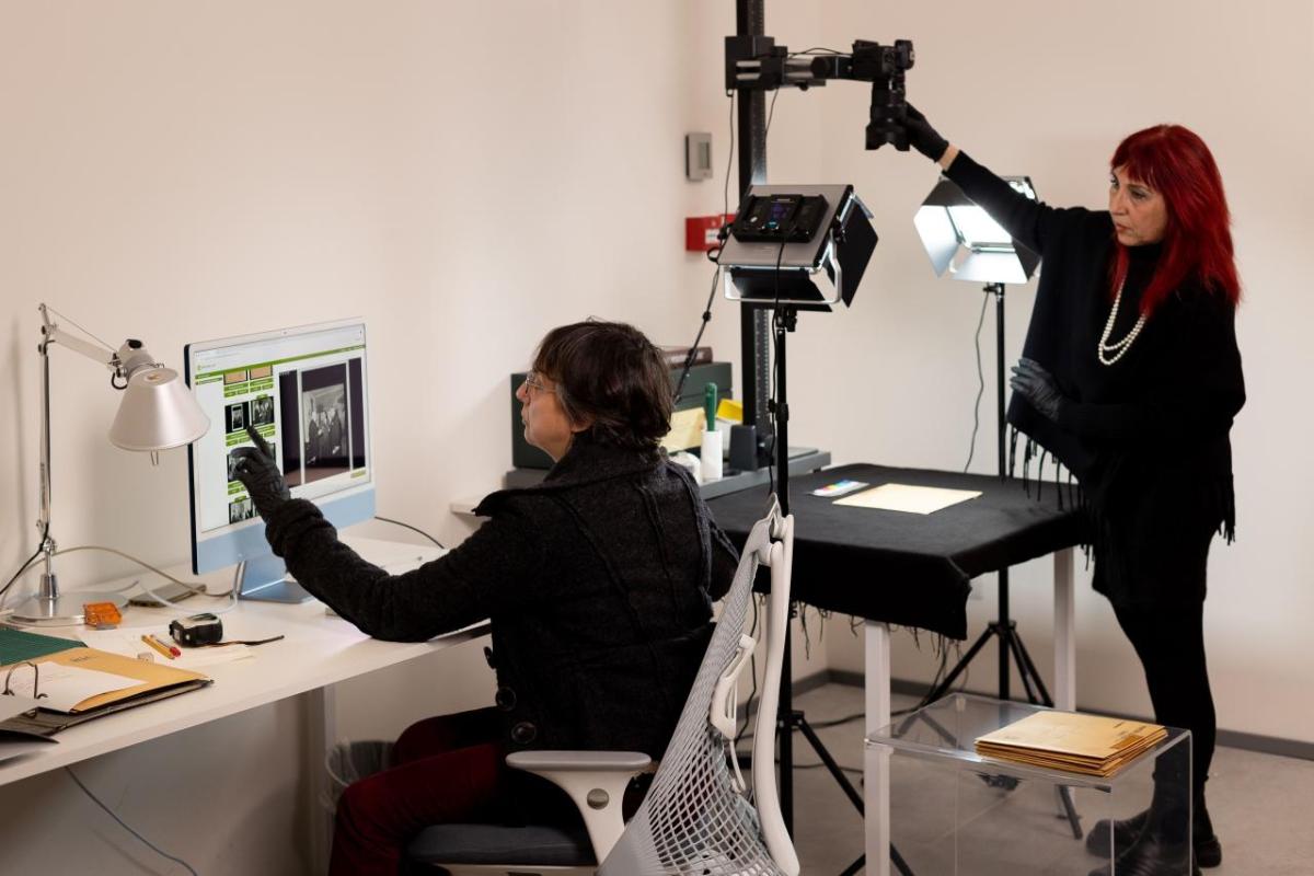 Two people in a room, one at a computer desk, pointing to an image on the screen. The other adjusting equipment above a photograph on a table.