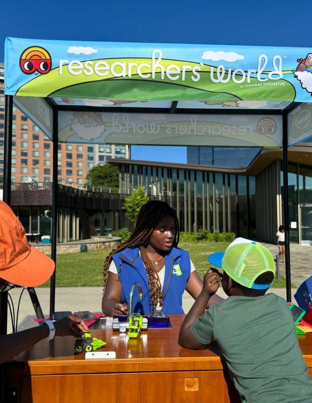 "Researchers' World" banner on an awning over a table with a person demonstrating to kids outside.