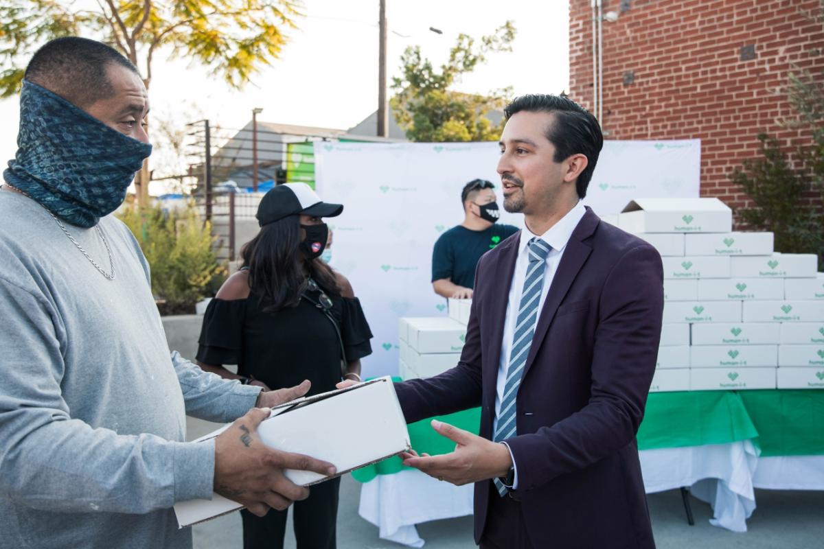 A man in a suit hands a Human-I-T box to a man in a mask