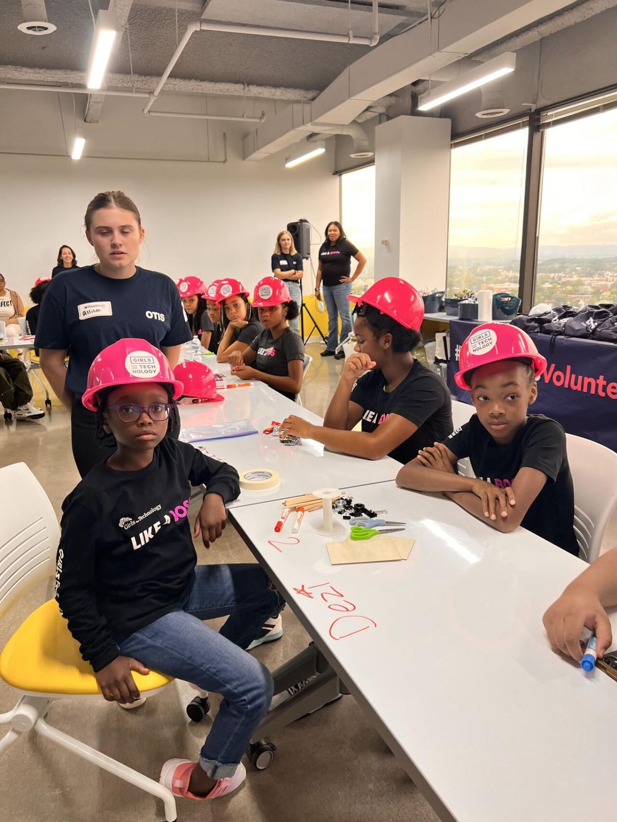 Children seated at long table wearing matching pink hard hats.