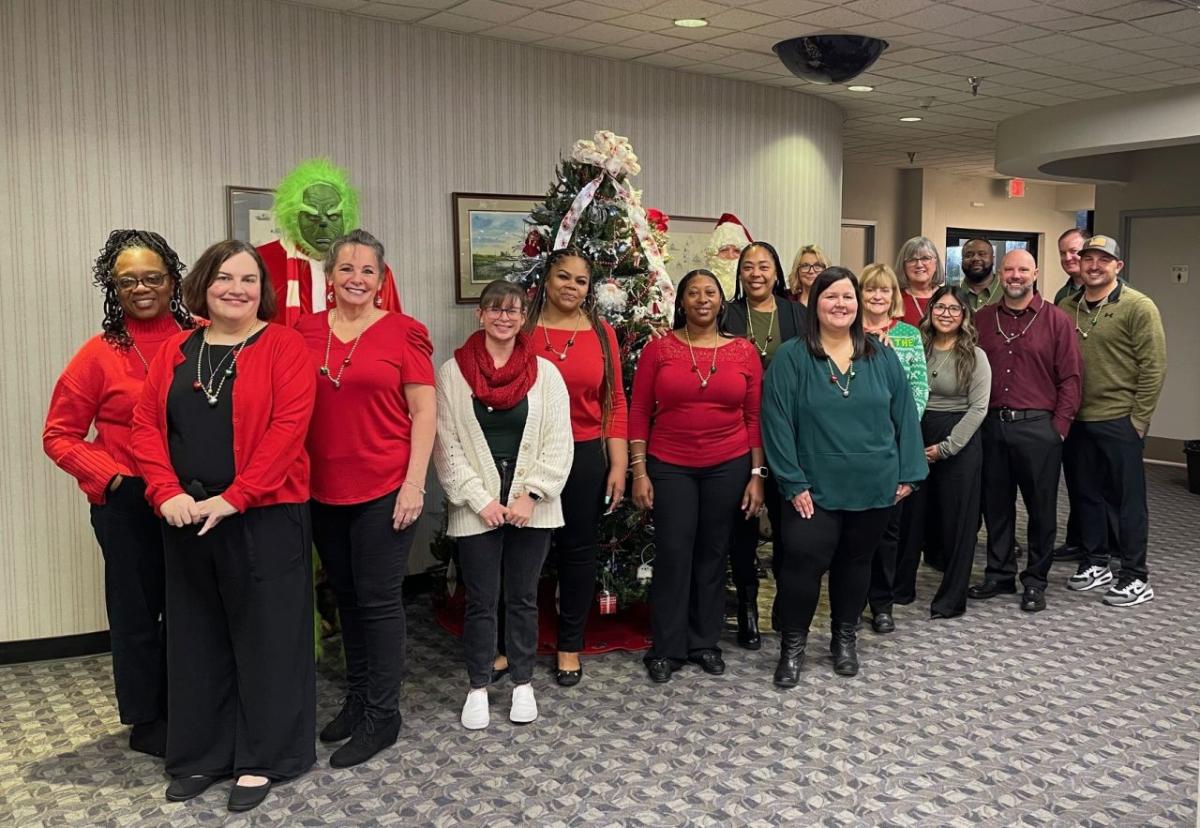 A group of volunteers posed in front of a chistmas tree. One dressed as the grinch behind them.