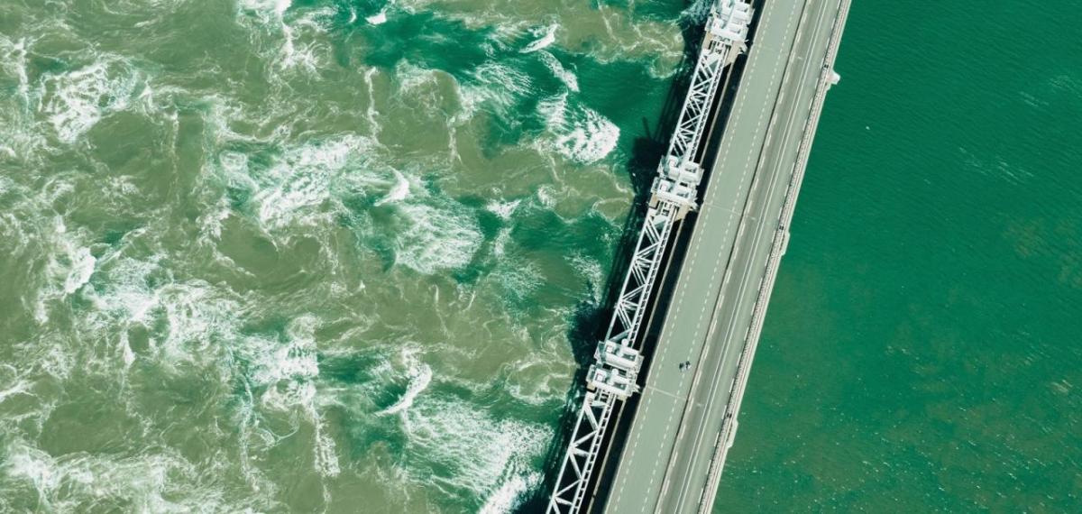 Aerial view of a large dam with green water on one side and cloudy green turbulent water on the other.