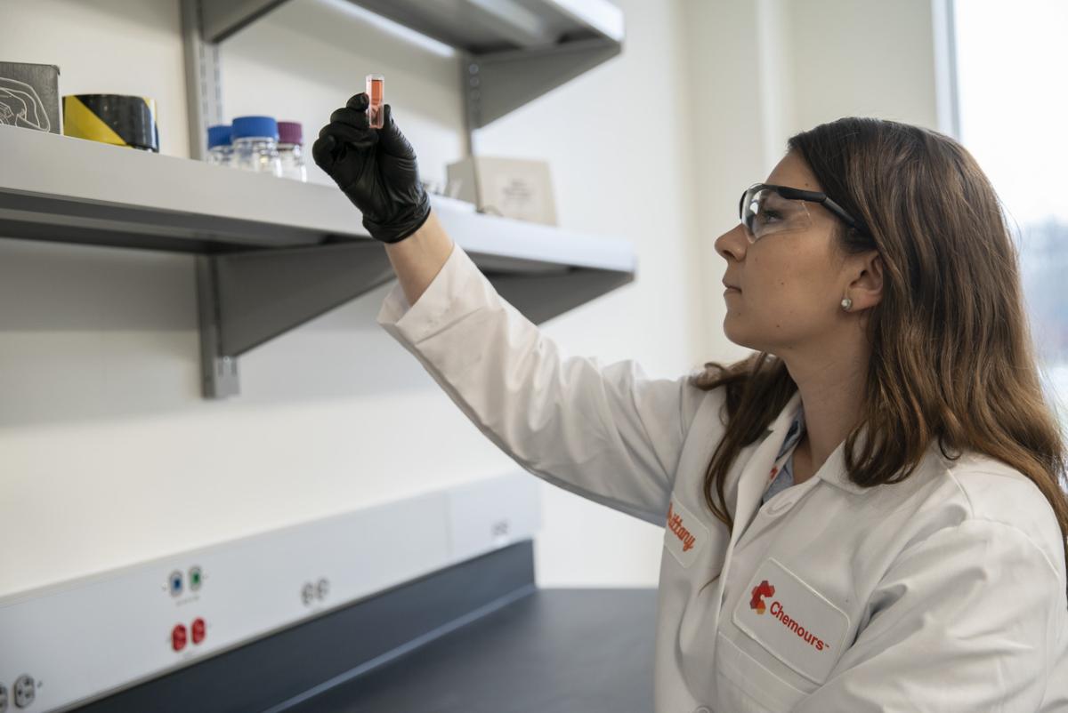 A person wearing protective lab coat, glasses and gloves, inspecting a fluid in a small tube in a laboratory setting.