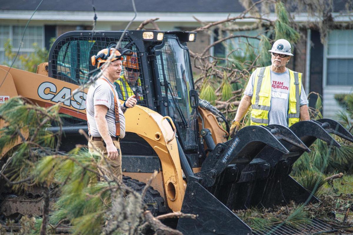 People standing next to a scooping types construction vehicle. Debris and branches surrounding.