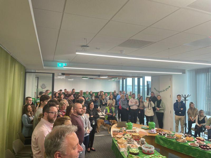 A room packed with people standing, an oval table with food on it. 