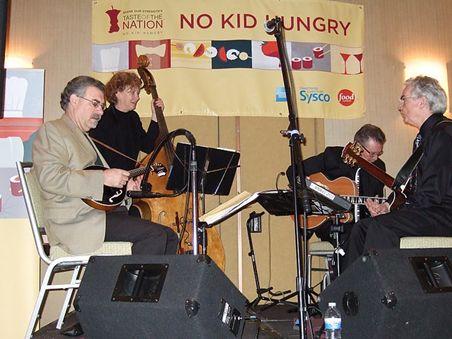 A small band playing on a stage. "No Kid Hungry" banner behind them.