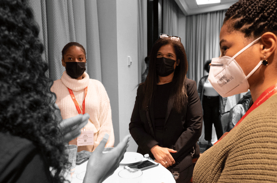 a group of people stand around a small round table, all wearing protective masks