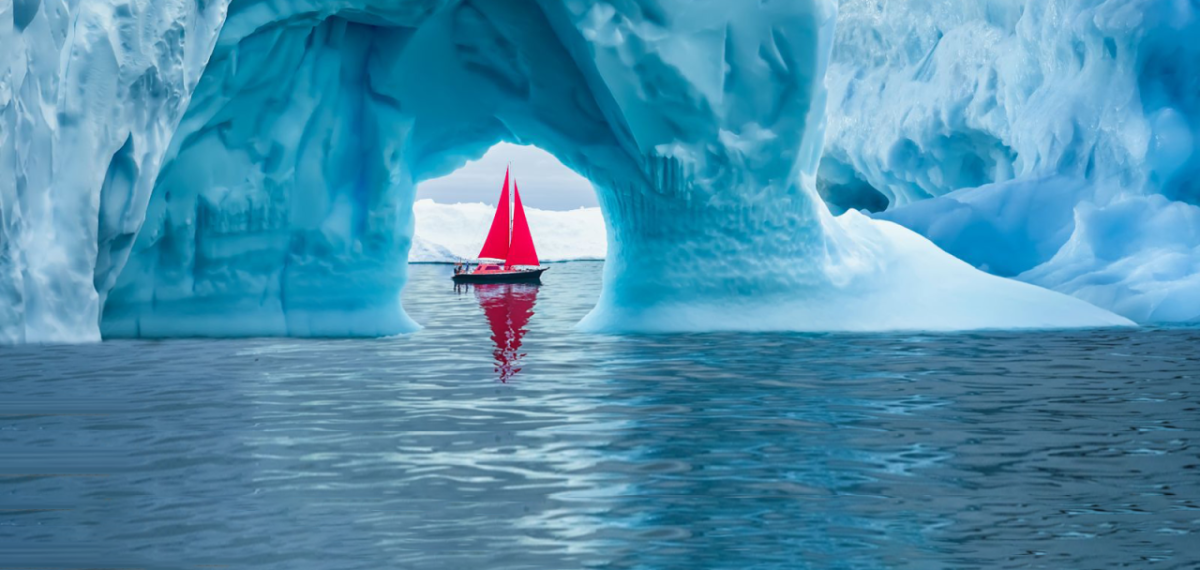 A boat with red sails framed in a small opening in an iceberg on a body of water.