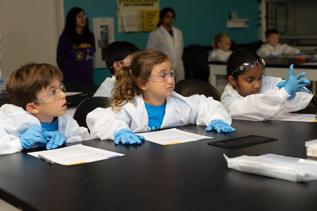 Three students in lab coats and goggles sitting in a science lab.