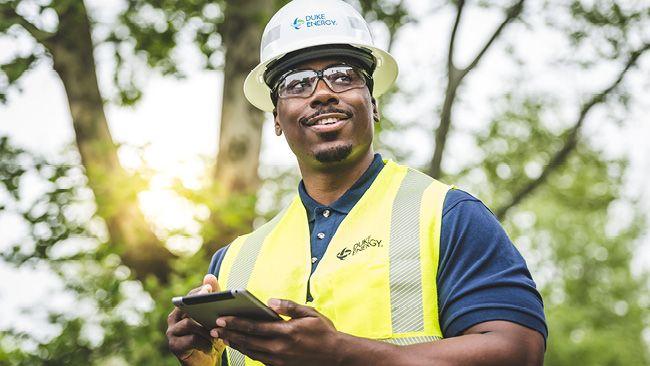 A person in safety hat and vest with a tablet in their hands, sun through the trees behind them.