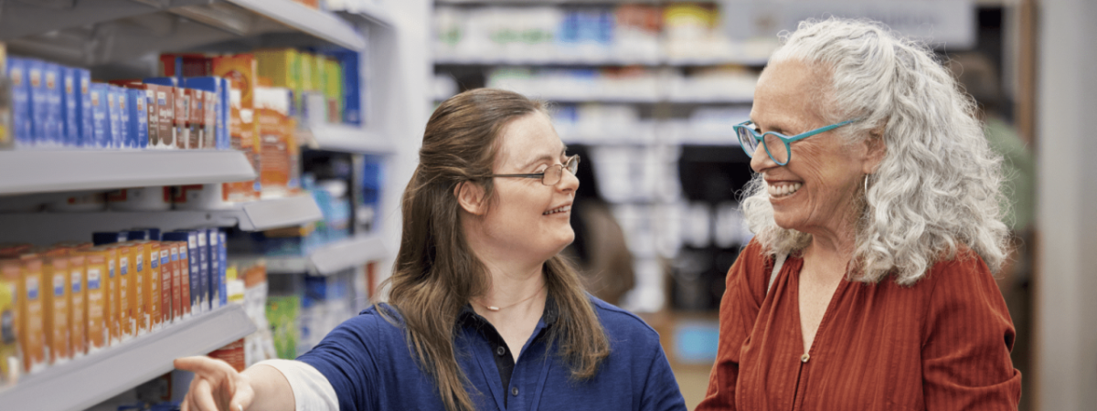 Two people smiling at each other in a pharmacy aisle, one points to an item on a shelf.