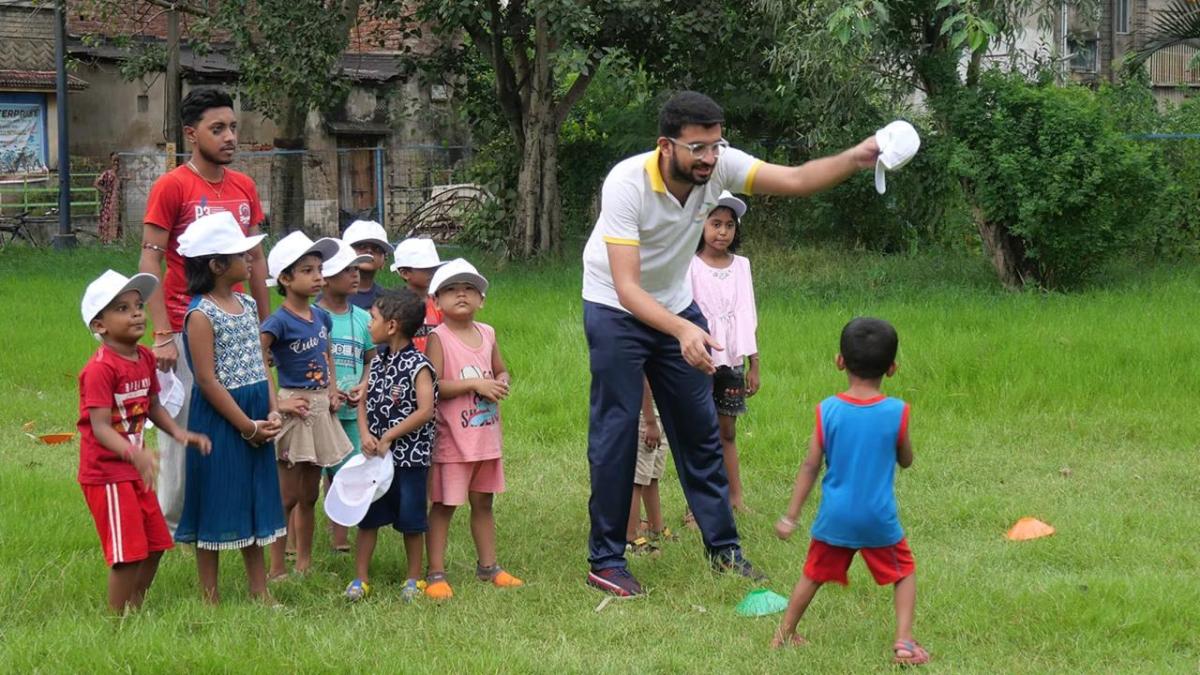 An adult shows a child a white hat, other children behind them in a group each with a matching white hat on.