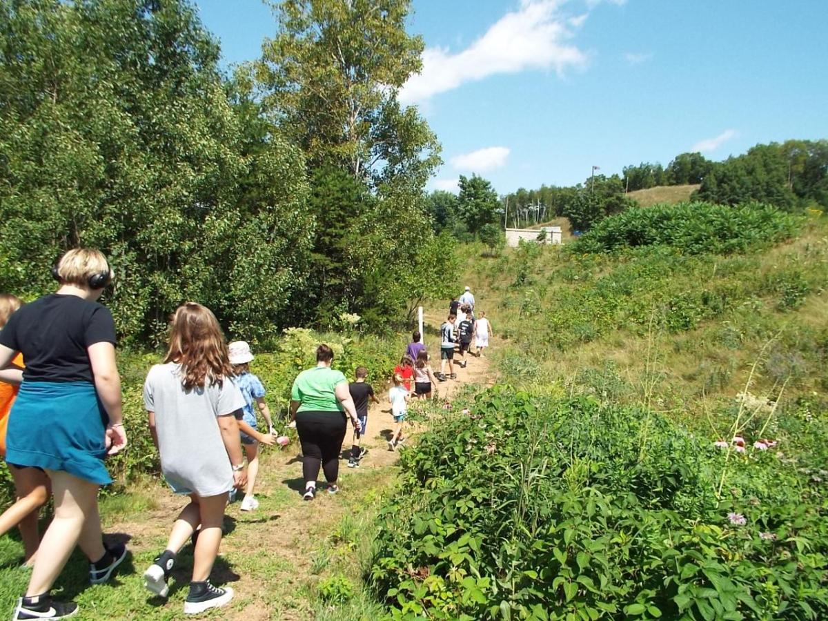 Kids and adults walk in a line along a narrow path in a lush area.