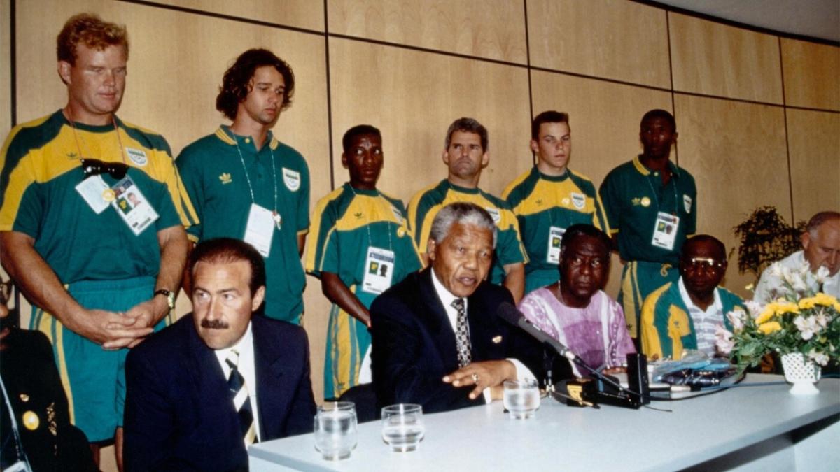 athletes in matching uniforms stand behind Nelson Mandela and other leaders seated at a long table