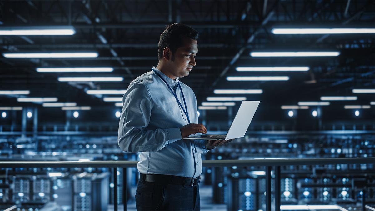 a person standing, working on a laptop, a warehouse of servers behind them