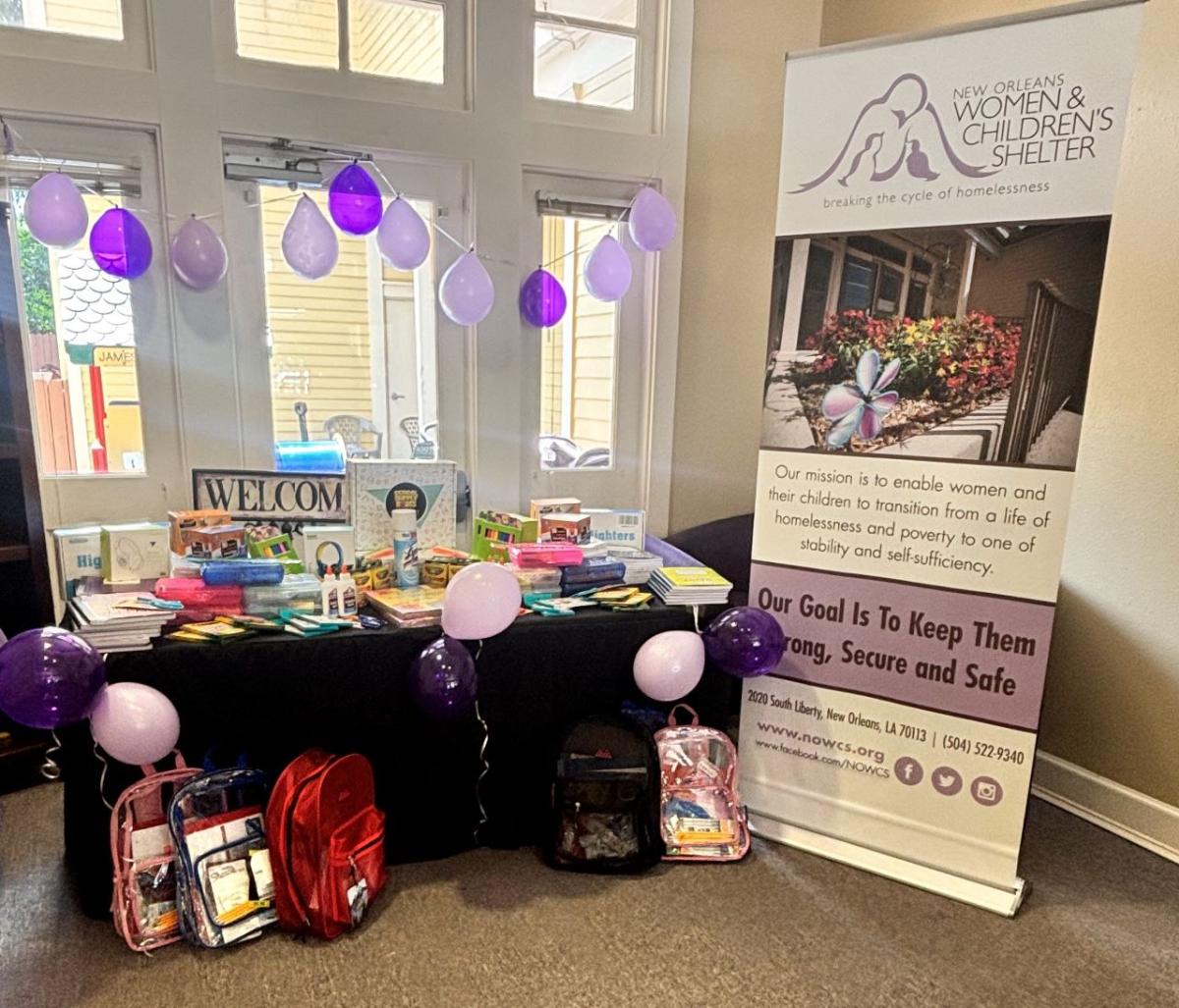 A table stacked with school supplies, with several new backpacks leaning against it