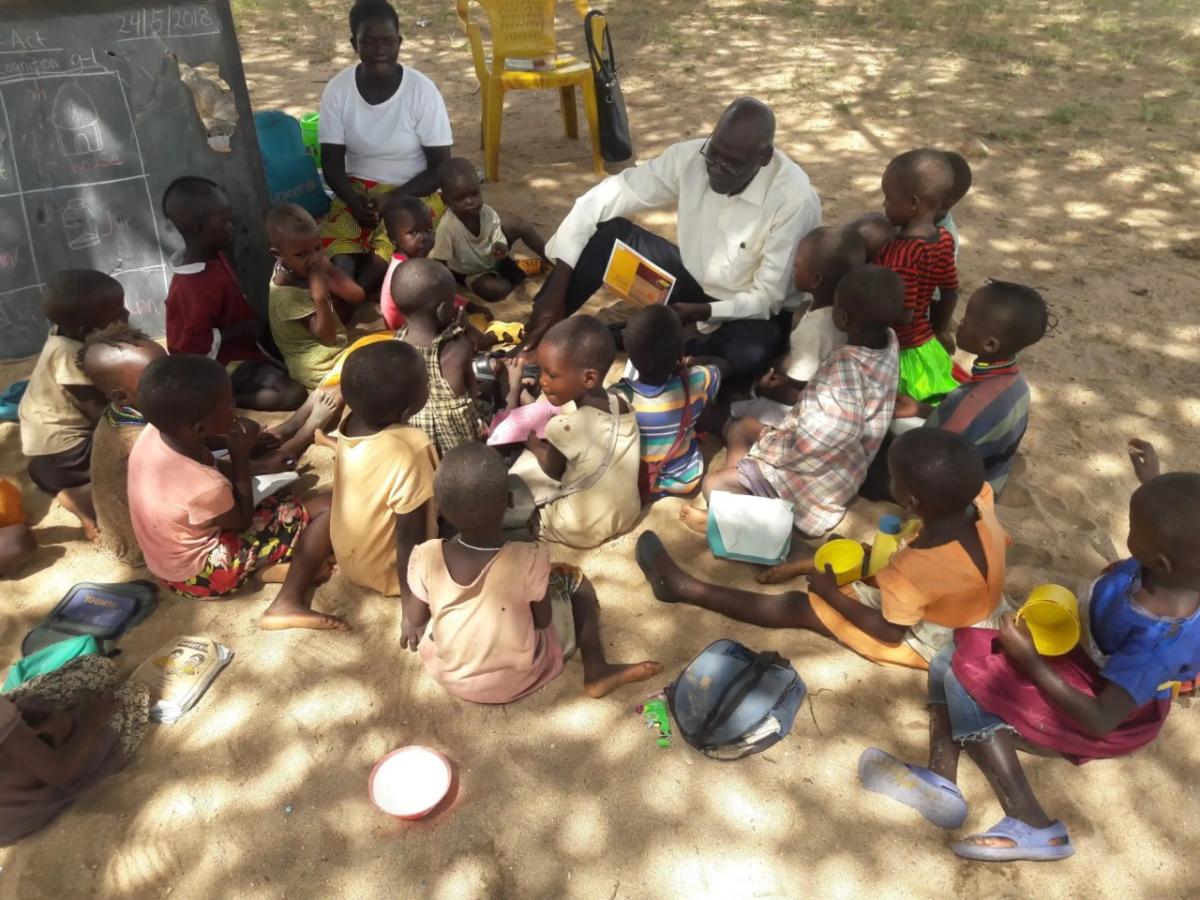 A group of children sitting on sandy ground, an adult showing them a picture