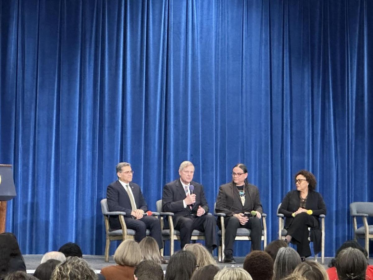 A panel of four seated on a stage in front of a room of people.