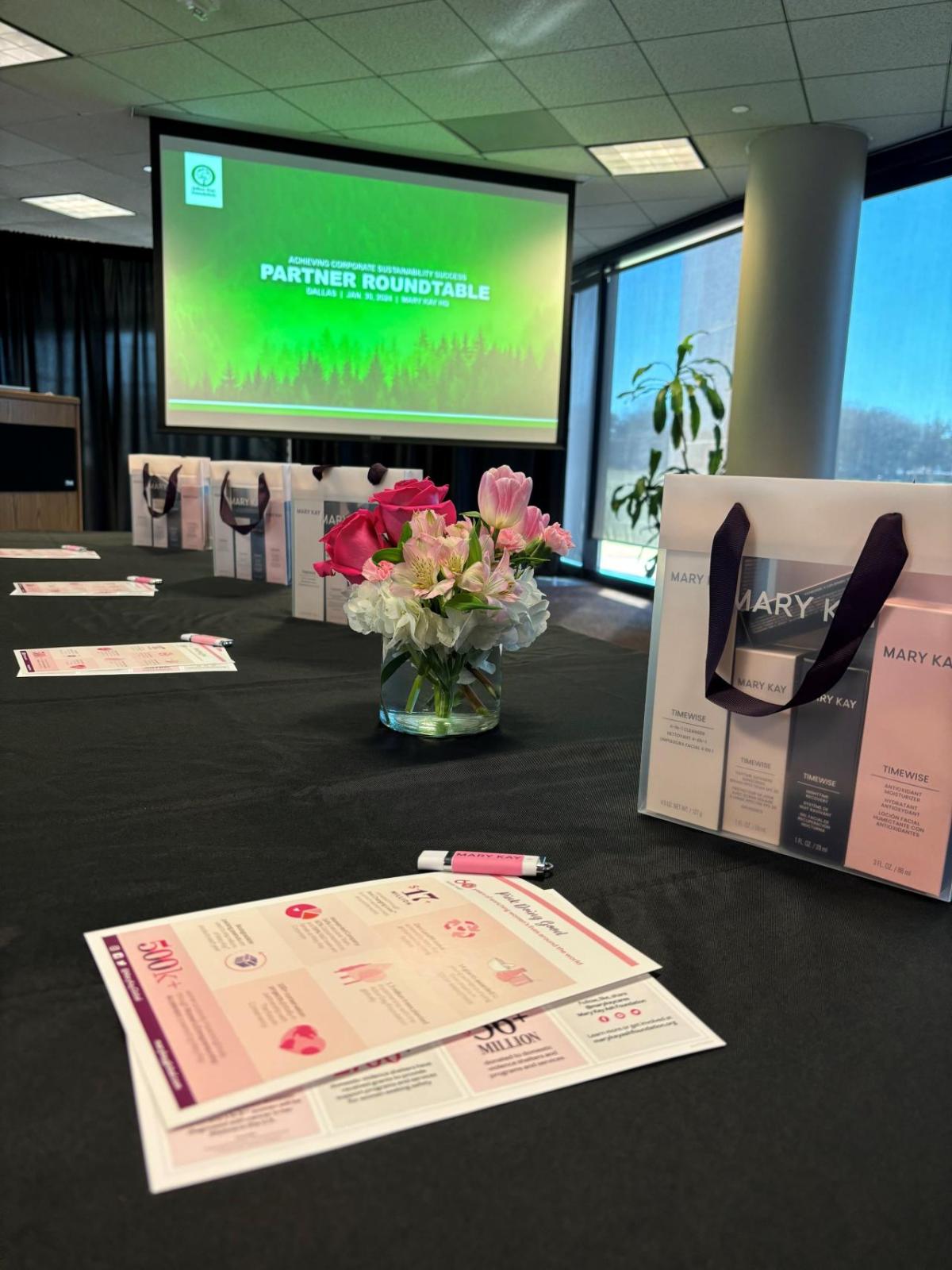 A table set with gift bags, papers and a pen. A small bouquet of flowers central. A pull-down screen in the background.